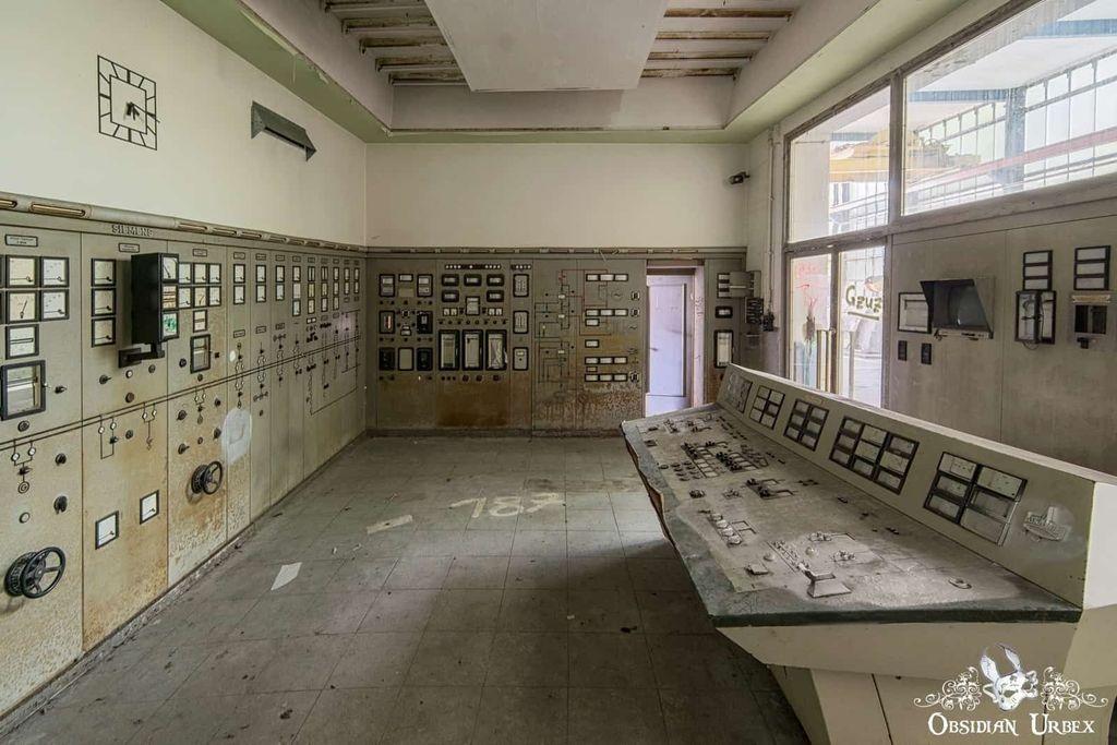 An abandoned control room with aging panels, numerous switches, dials, and meters, illuminated by light from large windows on the right.