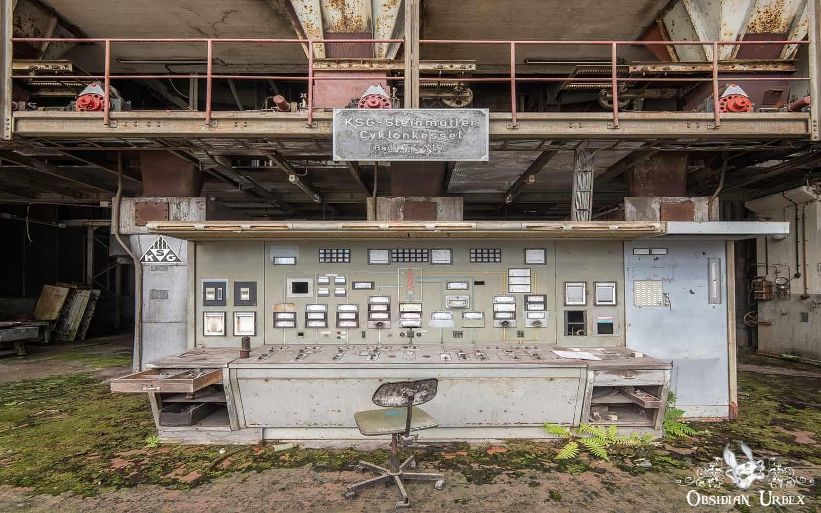 An old, rusted industrial control panel with numerous buttons and meters stands in an abandoned, decaying facility; a lone metal chair is in front.