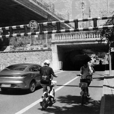 Ilford HP5 Plus 400 (6x6)

English Alt Text:
A black and white photo captures a city street running beneath a low overpass with a clearance sign reading “2.5m.” Two people ride bicycles or electric scooters—one holds an umbrella while moving. A car travels alongside them. The overpass has a stone wall and metal railings. In the distance, tall buildings and construction cranes suggest ongoing urban development. The monochrome tone adds a timeless, documentary feel. The scene reflects everyday ci…