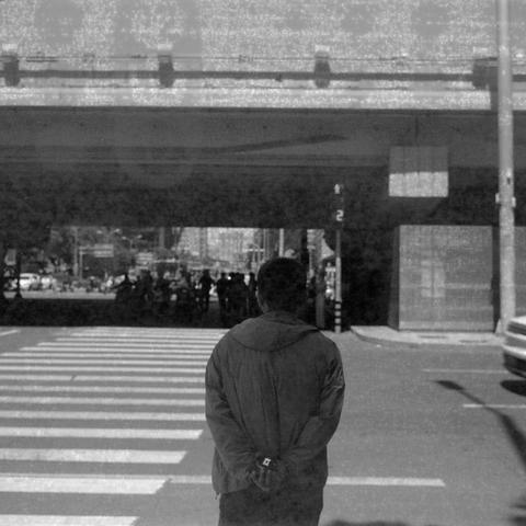 Ilford HP5 Plus 400 (6x6)

English Alt Text:
A contemplative black and white image shows a person standing at a crosswalk beneath an overpass. The person faces away from the camera, hands clasped behind their back, wearing a jacket. Shadows from the bridge create dramatic contrast on the pavement. In the background, several pedestrians and cars move through the urban setting. The photo evokes solitude and reflection, with strong light and shadow interplay.

中文替代文字：
这张黑白照片展现了一位行人站在高架桥下的斑马线上，背对镜头…