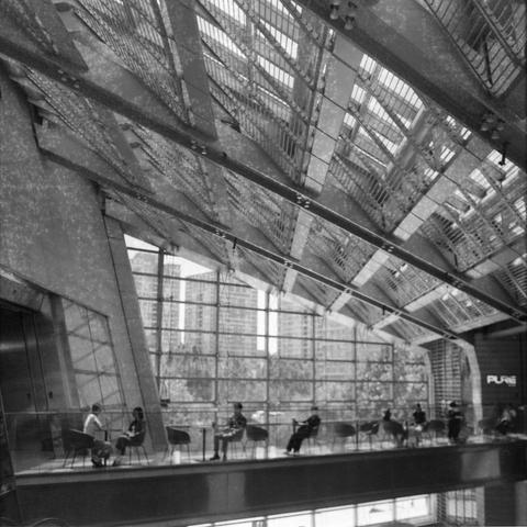 Ilford HP5 Plus 400 (6x6)

English Alt Text:
A black and white photo of a modern building interior with a high, angled glass ceiling supported by geometric beams. Natural light floods the space. On a mezzanine level, people sit at round tables, possibly in a café or lounge. The word “PURE” is visible on the right wall. Through the large windows, tall buildings suggest an urban setting. The architectural design and relaxed atmosphere contrast with the city outside.

中文替代文字：
这张黑白照片展示了一座现代建筑的内部，高耸…
