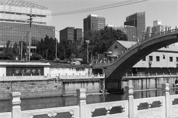 Kentmere Kentmere PAN 400 (FF)

English Alt Text:
A black-and-white photo of a stone bridge arching over a canal. The bridge has decorative railings and connects to a building on the right. The canal is lined with stone walls and calm water. A sign on the left. In the background, modern high-rises with glass facades rise above, blending tradition with contemporary architecture.

中文替代文字：
这是一张黑白照片，展示一座石桥横跨运河。桥上有装饰性栏杆，连接右侧建筑。运河两岸是石砌墙体，水面平静。左侧有一块标牌。背景是几栋现代玻璃高楼，传统与现代建筑在此交融。