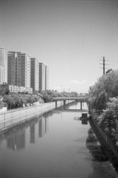 Kentmere Kentmere PAN 400 (FF)

English Alt Text:
A black-and-white photo of a calm urban canal. On the left, tall buildings rise above the water; on the right, leafy trees line the bank. A bridge spans the canal in the middle distance. Power lines and a utility pole are visible on the right. The water reflects the buildings and trees, creating a peaceful, symmetrical scene. The contrast between nature and urban development is emphasized by the monochrome tones.

中文替代文字：
这是一张黑白城市运河照片。左侧是高楼林立，右侧…