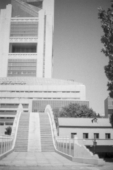 Kentmere Kentmere PAN 400 (FF)

English Alt Text:
A grayscale image of a modern cityscape. A curved pedestrian bridge with steps and railings dominates the foreground. Behind it, a striking high-rise with diagonal and horizontal structural elements and a glass facade towers above. To the right, a smaller building with a gabled roof and a tree adds contrast. The composition highlights architectural diversity and urban layering.

中文替代文字：
这是一张灰度城市景观照片。前景是一座弯曲的人行桥，带有台阶和栏杆。背景是一栋现代高楼，结构线条斜交纵横，外墙为玻璃材质…