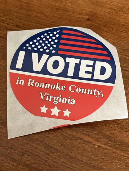 An "I Voted" sticker featuring a design with the American flag, stating "I VOTED in Roanoke County, Virginia" along with three stars at the bottom. The sticker is placed on a wooden surface.
