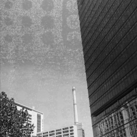 Ilford HP5 Plus 400 (6x6)

English Alt Text:
A monochrome photograph of a city scene with architectural contrasts. On the right, a tall building with a glass facade and grid-like pattern rises prominently. In the background, a mid-rise structure features a smokestack or chimney. A tree in the lower left corner partially obscures another building. The sky above has a textured overlay, possibly due to photographic effects. The image evokes a sense of industrial modernism and urban layering.

中文替代…