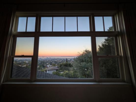Looking through a large multi-paned window eight minutes after sunrise we can see the sweep of the San Francisco Bay. The sky fades from bright peach at the horizon to pale blue above. We are high up, looking over houses and trees.