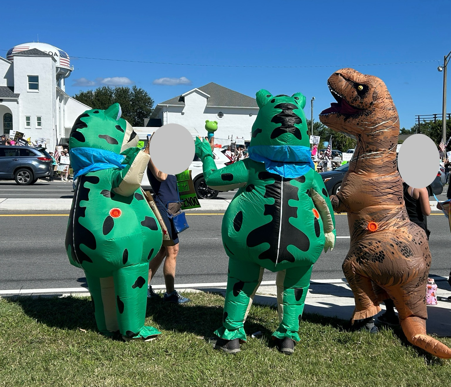 Two inflatable frogs standing next to an inflatable brown dinosaur, and a couple people not in costume.