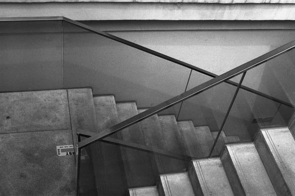 Kentmere Kentmere PAN 400 (FF)

English Alt Text: A black and white photograph of a modern staircase with a minimalist design. The steps are made of smooth concrete, and transparent glass panels serve as railings. A sleek metal handrail cuts diagonally across the image, intersecting with the geometric lines of the stairs and glass. The lighting casts soft shadows that emphasize the texture of the concrete and the clarity of the glass. The composition is clean and architectural, with a small lab…