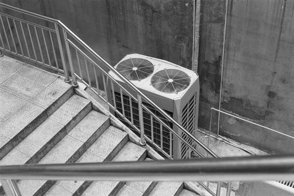 Kentmere Kentmere PAN 400 (FF)

English Alt Text: A black and white image of a stairwell with metal railings and tiled steps. Between the stair flights, a large air conditioning unit with two circular fans is wedged into a narrow space beside a worn concrete wall. The wall shows discoloration and age. The placement of the unit in such a confined area highlights the constraints of urban architecture and the creative use of space. The scene feels industrial and utilitarian, with a focus on functi…
