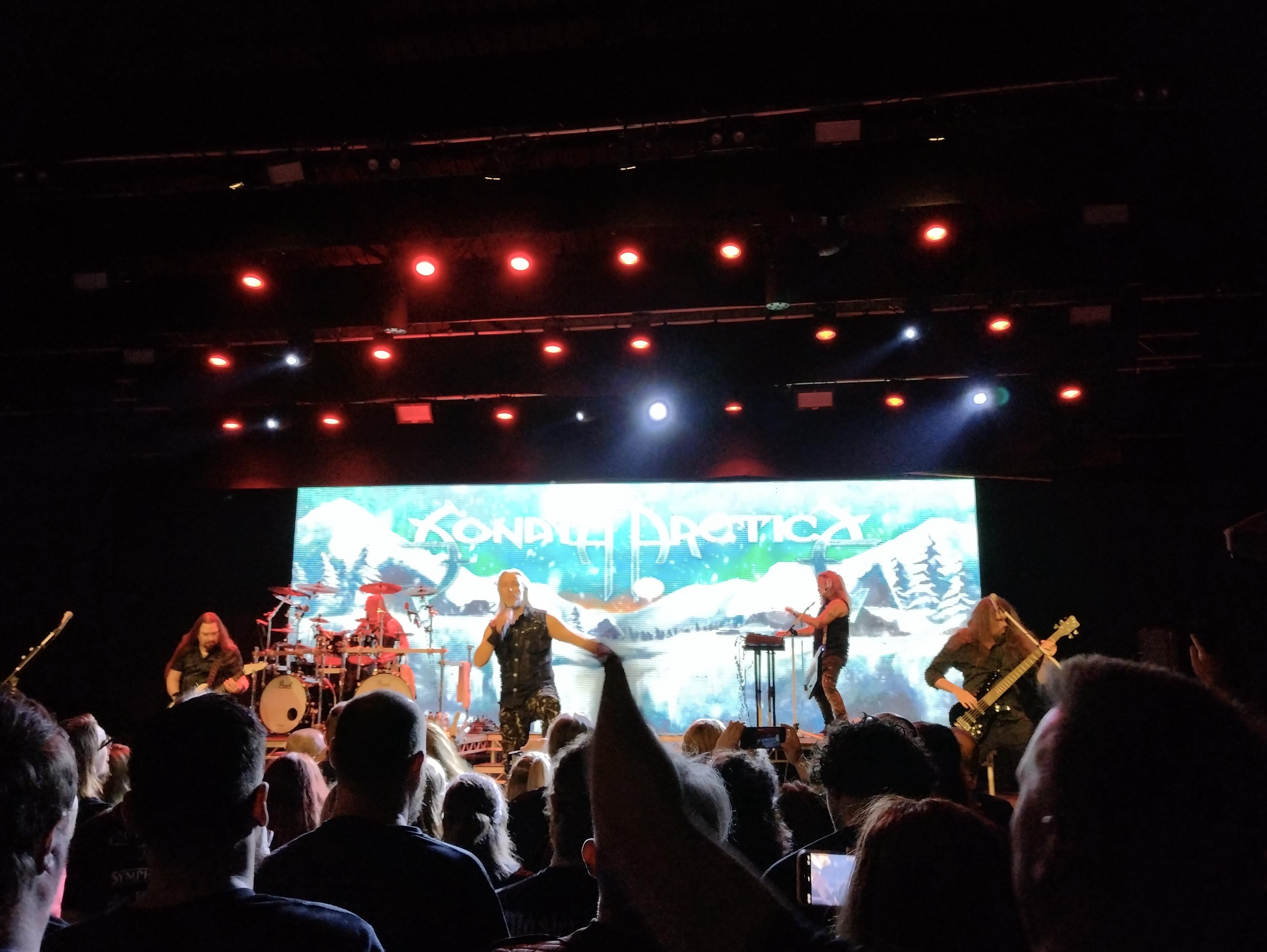 A band performs on stage with a backdrop featuring snowy mountains. The scene is illuminated by red stage lights, and the audience is visible in the foreground, enjoying the show.