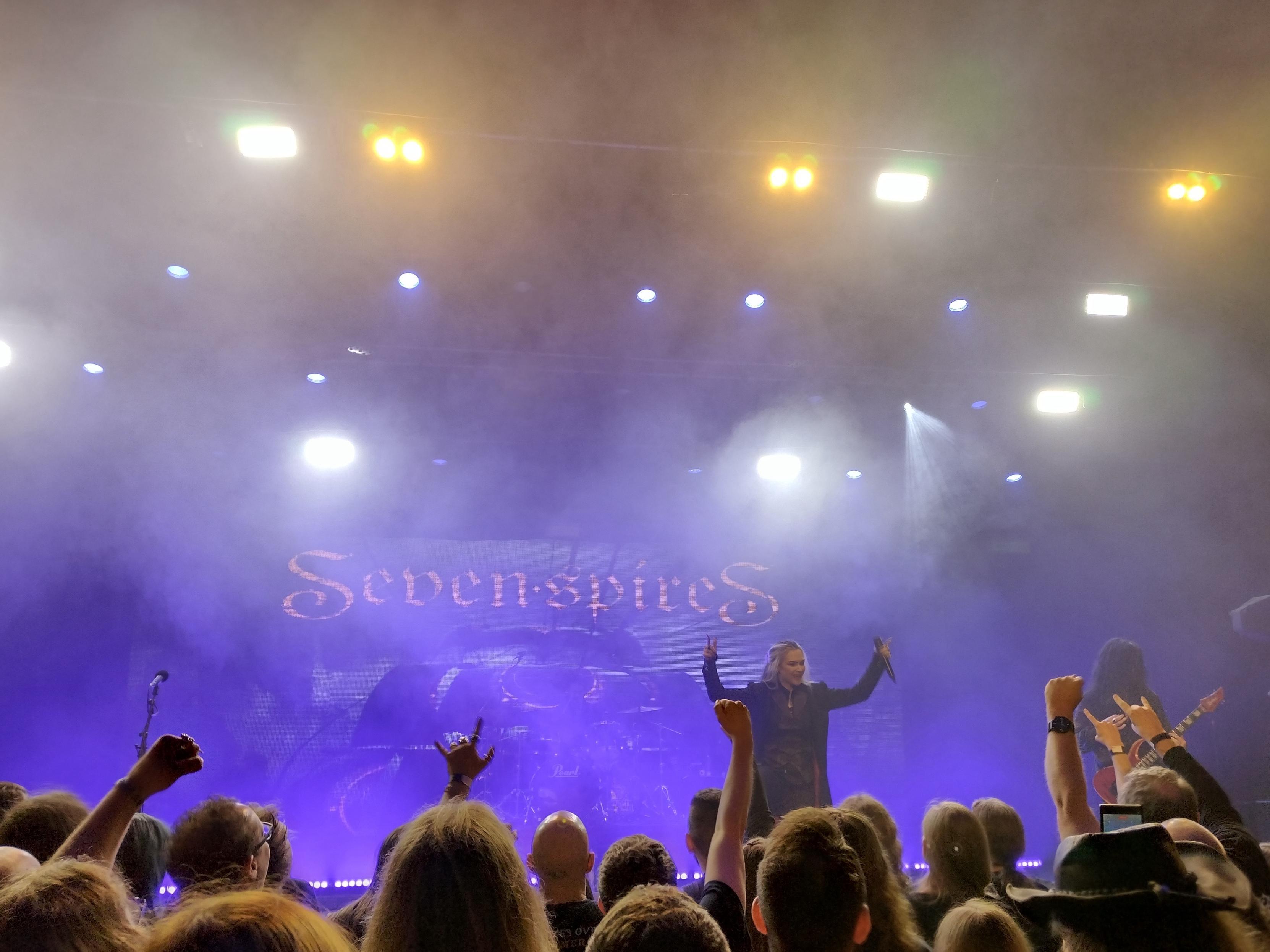 A live concert scene featuring the band Seven Spires on stage. The performance is surrounded by fog and colorful stage lighting, with an enthusiastic crowd visible in the foreground, some people with hands raised in excitement.