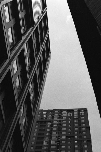 Kentmere Kentmere PAN 400 (FF)

English Alt Text:
A black-and-white photograph taken from a low angle, looking up at three towering modern buildings. The buildings have grid-like facades with rows of windows. Two structures frame the left and right sides of the image, while a third building rises in the center background. The sky is visible above, creating a stark contrast with the dark tones of the buildings. The perspective emphasizes verticality and scale, evoking a sense of urban grandeur a…