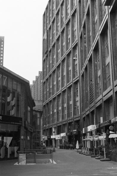 Kentmere Kentmere PAN 400 (FF)

English Alt Text:
A black-and-white street-level view of a narrow pedestrian street flanked by tall modern buildings. The facades feature grid-like windows and balconies. On the right, storefronts with signage, awnings, and outdoor seating suggest cafes or restaurants. On the left, a building with large glass windows displays partial signage reading “ITAL” and “GN.” A glass-covered entrance in the foreground may lead underground. The scene is quiet, emphasizing u…