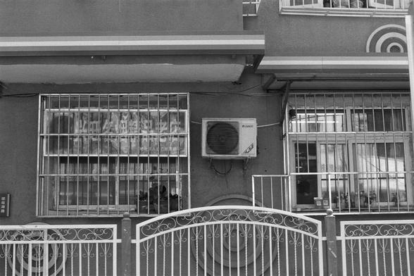 Kentmere Kentmere PAN 400 (FF)

English Alt Text:
A black-and-white photo of a building with barred windows and a decorative facade featuring horizontal stripes and circular patterns. An air conditioning unit is mounted between two windows. One window has partially obscured Chinese signage. A metal gate with ornate designs stands in front of the entrance. The image evokes a vintage or documentary feel, highlighting architectural details and urban texture.

中文替代文本：
这是一张黑白照片，展示一栋建筑，窗户装有铁栏杆，外墙装饰有横…