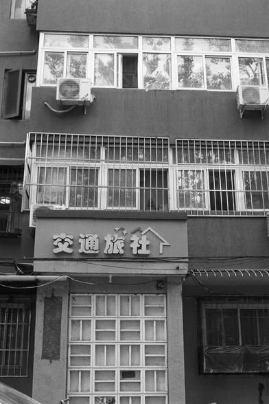 Kentmere Kentmere PAN 400 (FF)

English Alt Text:
A black-and-white image of a multi-story commercial building. The lower level features a sign with Chinese characters reading “交通旅行社” (Transportation Travel Agency). Above are barred windows and air conditioning units. The upper floors have enclosed balconies with glass windows reflecting trees and sky. The building’s facade suggests a mix of residential and business use in an urban Chinese setting.

中文替代文本：
这是一张黑白照片，展示一栋多层商业建筑。底层有一块中文招牌，写着“交通旅行…