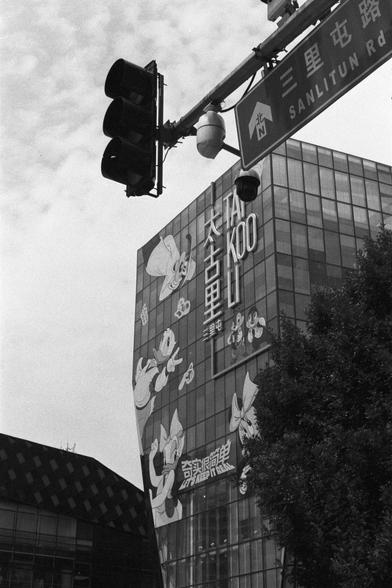 Kentmere Kentmere PAN 400 (FF)

English Alt Text:
A black-and-white photo of a modern glass building in Beijing’s Sanlitun area. The facade displays large cartoon characters and bilingual text: “TAIKOO LI” in English and “太古里” in Chinese. Below, the phrase “奇境探险” translates to “Adventure in Wonderland.” A traffic light and street sign reading “三里屯路 / SANLITUN Rd” are visible in the foreground. The image captures the vibrant, commercial atmosphere of a popular shopping and lifestyle district, bl…