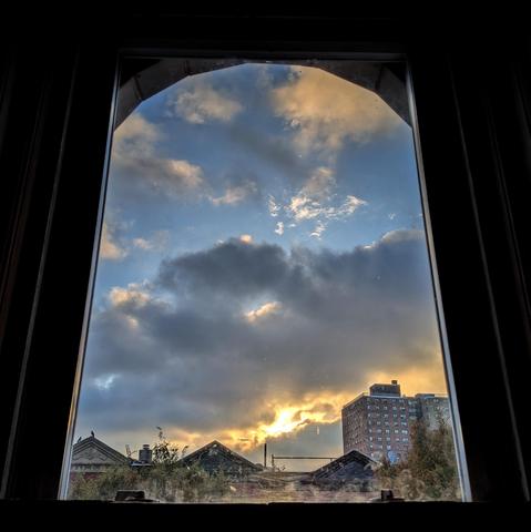 Looking through an arched window forty minutes after sunrise the blue sky is filled with large chunky gray and white clouds. The sun is breaking through in the lower sky, creating yellow highlights on the underside of the clouds. Pointed roofs of Harlem brownstones are silhouetted across the street, and a taller apartment building can be seen in the distance. The tops of two trees are on the bottom and right. The window is grimy on the bottom.