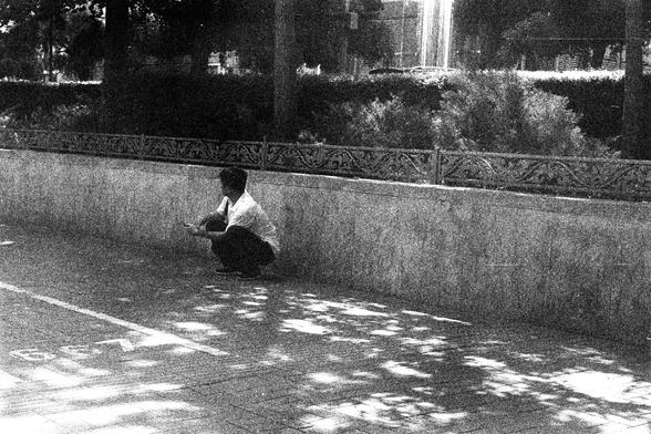 ORWO P400 (FF)

English Alt Text:
A grainy black-and-white photo shows a person squatting on a paved sidewalk beside a low concrete wall topped with decorative metal railings. The area is shaded by leafy trees, casting dappled light and shadow patterns on the ground. In the background, a water fountain sprays upward, surrounded by greenery. Parked cars and buildings are partially visible through the trees. The scene feels quiet and contemplative, with a vintage film-like texture. A white markin…