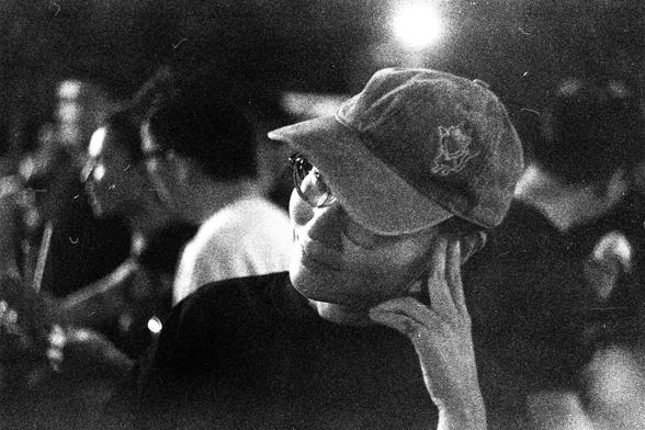 ORWO P400 (FF)

English Alt Text:
A grainy black-and-white image shows a person in the foreground wearing a cap with an emblem and glasses. They rest their head on their hand in a contemplative pose. The background is softly blurred, with several people visible, suggesting a social gathering. A bright light source above adds contrast and texture. The photo evokes a quiet moment of reflection amid a communal setting.

中文替代文本：
这是一张颗粒感强烈的黑白照片，前景中一人戴着有徽章的帽子和眼镜，用手托着头，姿态沉思。背景模糊，有几个人影，暗示这是一个社交场合。上方有强光…