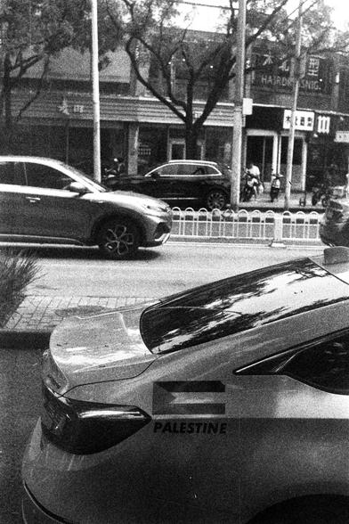ORWO P400 (FF)

English Alt Text:
A black-and-white street scene shows a parked car in the foreground with the word “PALESTINE” and a flag decal on its rear side. The car is near a sidewalk, and other vehicles are visible on the road, including a moving SUV. Across the street, storefronts display signage in Chinese characters. A group of people stands near one of the entrances. Trees, streetlights, and a pedestrian barrier add to the urban atmosphere.

中文替代文字：
这是一张黑白城市街景照片，前景是一辆停在路边的汽车，车尾贴有“PAL…