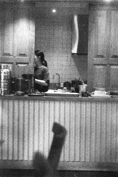 ORWO P400 (FF)

English Alt Text:
A grainy black-and-white photo shows a kitchen or food prep area. A person with a ponytail stands behind a counter, partially hidden by a cabinet door, appearing to prepare food or drinks. The counter holds a large metal container, cups, and a coffee machine. The background includes wooden cabinets and tiled walls.

中文替代文字：
这是一张黑白颗粒感照片，拍摄于厨房或食物准备区。一位扎着马尾的人站在柜台后方，部分被橱柜门遮挡，似乎正在准备食物或饮品。柜台上有一个大金属容器、杯子和咖啡机。背景是木质橱柜和瓷砖墙面。