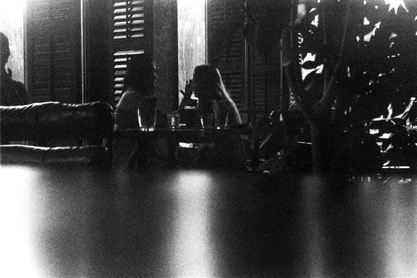 ORWO P400 (FF)

English Alt Text:
A grainy black-and-white photograph captures a quiet moment in a dimly lit café or restaurant. Two people sit across from each other at a small table, engaged in conversation. The background features shuttered windows and scattered furniture, including empty chairs and tables. A leafy plant partially obscures the right side of the image. The foreground is blurred, possibly due to a reflective surface or intentional photographic effect, adding depth and intimacy…