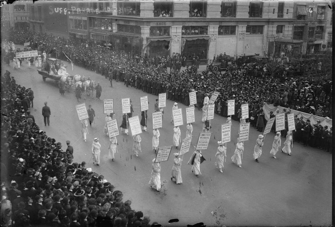 large crowds watch the parade. The front has 24 women all in white and carrying banners. Everyone is white.