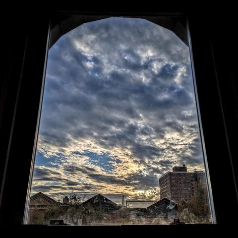 Looking through an arched window an hour after sunrise the blue sky is filled with large chunky blue-gray clouds in the closer high sky, and scattered white and gray clouds backlit by the sun closer to the horizon. Pointed roofs of Harlem brownstones are silhouetted across the street, and a taller apartment building can be seen in the distance. The window is grimy on the bottom.