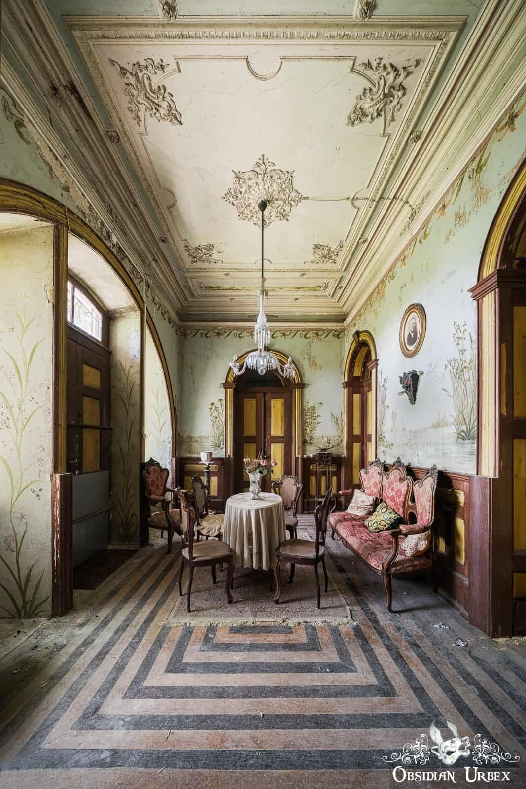 The room appears to be a dining or sitting area, featuring ornate, faded wallpaper with a floral pattern and a geometric-patterned floor. A chandelier hangs from the detailed, decorative ceiling, illuminating a round table with a tablecloth and four chairs in the center of the room. An upholstered settee sits against the right wall.