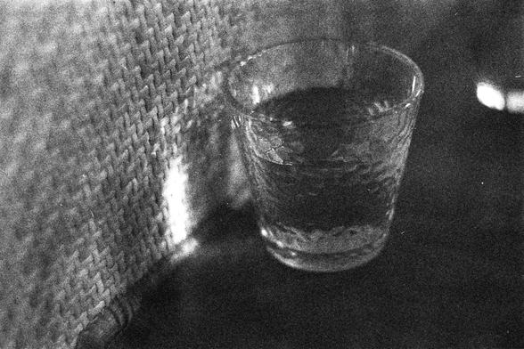 ORWO P400 (FF)

English Alt Text:
A black-and-white close-up of a textured glass partially filled with a dark liquid, placed on a dark surface. Behind it is a woven or patterned wall panel, illuminated by light that casts reflections and shadows. The grainy texture and interplay of light and surfaces create a moody, intimate composition.

中文替代文字：
这是一张黑白特写照片，画面是一只表面有纹理的玻璃杯，杯中装有部分深色液体，放在深色表面上。背景是一块编织或有图案的墙板，被光照亮，形成反射和阴影。图像颗粒感强，光影与材质的互动营造出一种私密而感性的氛围。