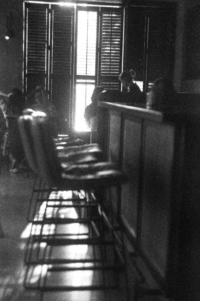 ORWO P400 (FF)

English Alt Text:
A black-and-white photo of a dimly lit bar or restaurant interior. In the foreground, a row of cushioned bar stools with metal legs lines a wooden-paneled counter. Sunlight streams through tall, shuttered windows in the background, casting dramatic shadows across the floor and furniture. Several people sit and converse in the distance, partially obscured by light and shadow. The grainy texture adds a nostalgic, moody atmosphere.

中文替代文字：
这是一张黑白照片，拍摄的是一个昏暗的酒吧或餐厅…
