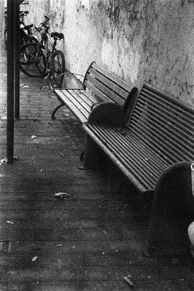 ORWO P400 (FF)

English Alt Text:
A monochrome photograph of a sidewalk scene. Two empty metal benches with slatted seats sit against a textured, worn wall. Several bicycles are parked nearby, some leaning against the wall. A vertical pole stands near the center. Leaves and debris scatter across the ground, suggesting neglect. The grainy texture and composition evoke a sense of quiet urban decay.

中文替代文字：
这是一张黑白人行道场景照片。两张空的金属长椅靠着一面有纹理且破旧的墙壁。几辆自行车停在附近，有些靠在墙上。画面中央有一根垂直的杆子。地面上散落着树叶和杂物，透露出些许荒废感。颗粒感…