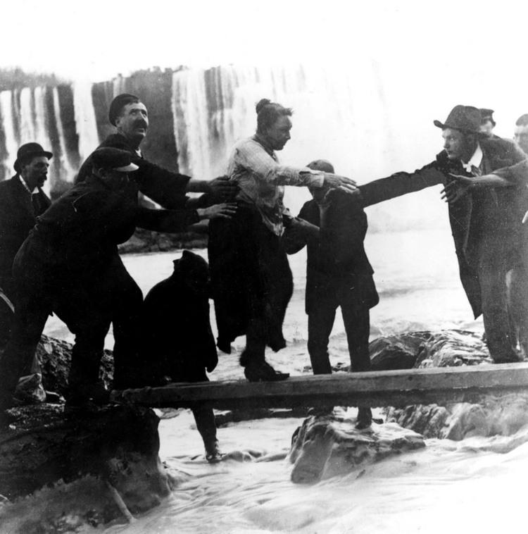 Annie Edson Taylor being helped ashore along a plank, having survived the falls. She is a white woman in a blouse and skirts.