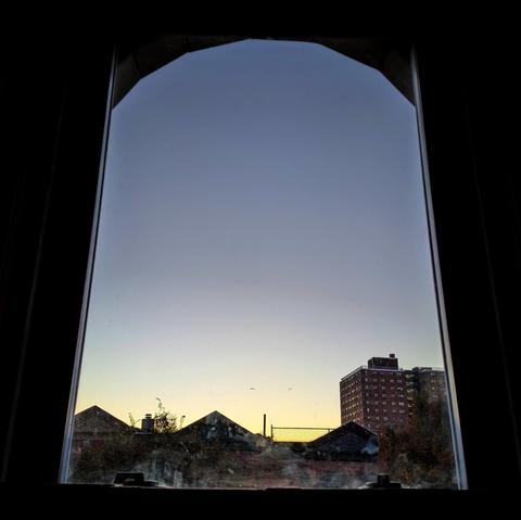 Looking through an arched window at the moment of sunrise the cloudless sky fades from pale yellow at the horizon to robin-egg blue up above. Pointed roofs of Harlem brownstones are silhouetted across the street, and a taller apartment building can be seen in the distance. 