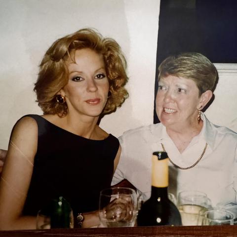 Photo of me in a black bateau neck dress with shoulder length red hair sitting at a restaurant table with an older woman with short grey & blonde hair & wearing a white blouse and earrings, who’s looking at me & is about to speak. There are glasses and a bottle of wine in the foreground.