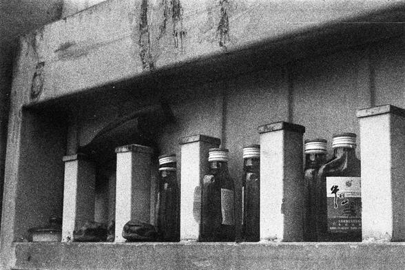 ORWO P400 (FF)

English Alt Text:
A black-and-white image shows a shelf with several bottles, each in its own partition. The bottles have screw caps and labels; one features Chinese characters and a cow image, possibly indicating a dairy product. The shelf surface is worn and grimy. The juxtaposition of everyday items creates tension and mystery.

中文替代文本：
这是一张黑白照片，画面中一个隔间式架子上排列着几瓶饮品。瓶子有旋盖和标签，其中一个标签上有中文和牛的图像，可能是乳制品。瓶子之间隐约可见一把手枪，被隔板部分遮挡。架子表面磨损严重，显得肮脏。日常物品与武器的并置营造出紧张和神秘的氛围。