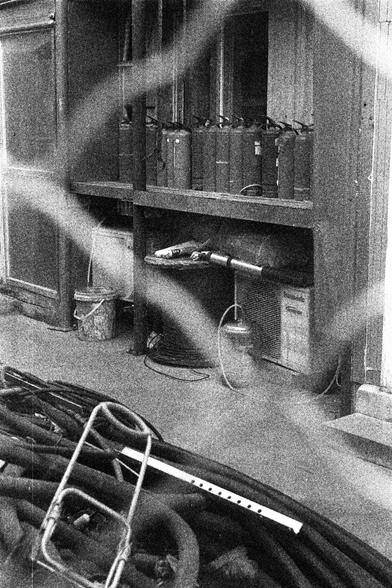 ORWO P400 (FF)

English Alt Text:
A cluttered storage area is shown in black and white. Red metal containers, possibly for fuel or oil, are lined on a shelf. Below are coiled hoses, a bucket, and miscellaneous tools. A wire mesh partially obscures the foreground. On the ground, more cables and a white measuring stick are visible. The scene suggests a workshop or industrial setting, with a gritty, utilitarian atmosphere.

中文替代文本：
这是一张黑白照片，展示一个杂乱的储物区。红色金属容器（可能用于燃料或机油）排列在架子上。下方有盘绕的软管、水桶和其他工具。前景被铁丝…