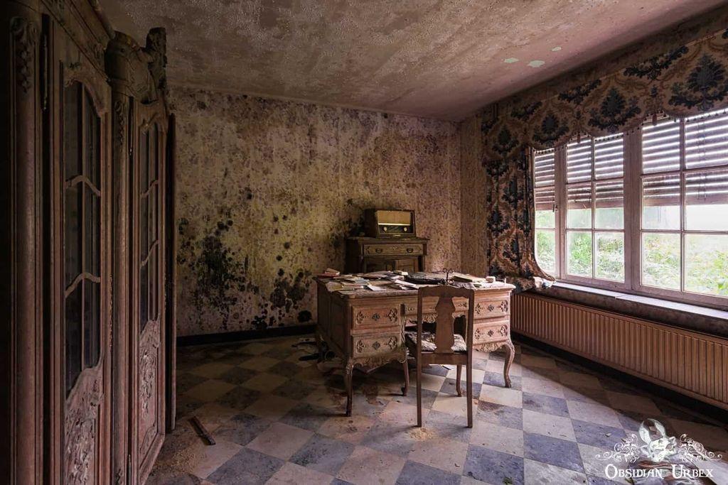 Abandoned room with moldy walls, dusty checkered floor, two ornate wooden desks covered in papers, large window with blinds, and vintage armoire.