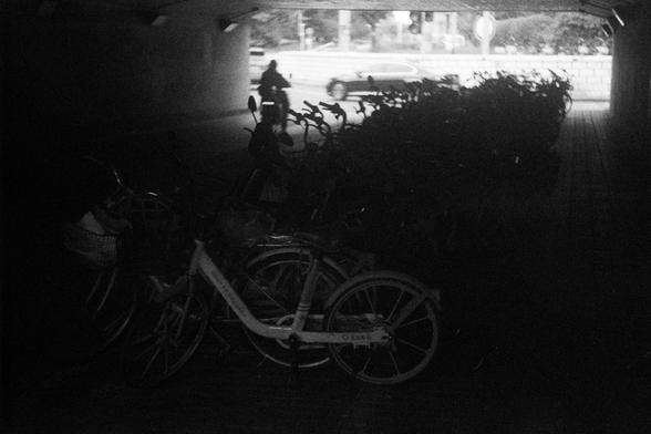 ORWO P400 (FF)

English Alt Text: A dimly lit tunnel filled with rows of parked bicycles. The tunnel’s interior is dark, with light pouring in from the far exit, creating a dramatic contrast. A person is silhouetted against the light, walking or cycling toward the exit. Outside, a car is visible on the road, along with greenery and a stone wall. The image captures an urban scene emphasizing bicycle use and the interplay of light and shadow.

中文替代文字：
一个昏暗的隧道里停满了自行车，排列整齐。隧道尽头透进明亮的光线，形成强烈的明暗对比。一名行…