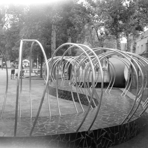 Ilford HP5 Plus 400 (6x6)

English Alt Text: A black-and-white image of a playground featuring a tunnel-like structure made of curved metal bars. The bars form looping arches over a tiled platform, inviting children to crawl or walk through. In the background, trees, benches, and a few people—including a child and an adult—add life to the scene. Urban buildings peek through the trees, hinting at a city setting. The structure’s artistic design and rhythmic curves create a playful yet sculptural …