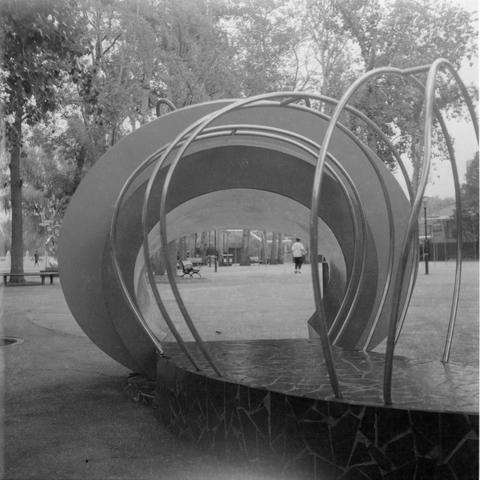 Ilford HP5 Plus 400 (6x6)

English Alt Text: A black-and-white image of a public park featuring a modern sculpture or playground structure. The foreground shows a circular base made of dark stone-like material, with curved metal rods arching overhead to form a dome-like shape. A large circular element behind it may be part of the same installation. Trees, benches, and people in the background suggest a peaceful, communal space. The abstract design adds a sense of creativity and modernity to the…