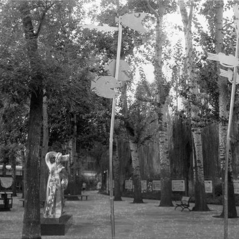 Ilford HP5 Plus 400 (6x6)

English Alt Text: A tranquil black-and-white photo of a park walkway lined with tall trees and artistic installations. A prominent sculpture of a human figure with raised arms stands in the foreground, made of reflective or metallic material. Along the path, tall poles display fish-shaped decorations, possibly wind vanes or art pieces. Benches and framed posters suggest the area may host exhibitions. The blend of nature and art creates a peaceful, contemplative atmosp…