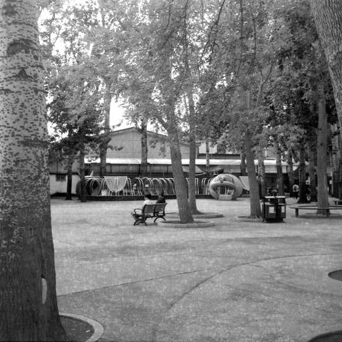 Ilford HP5 Plus 400 (6x6)

🖼️ Image 1: Park with circular structures and benches

English Alt Text: A black-and-white photo of a public park with tall trees and a paved surface. In the background, a building features a row of circular structures that resemble amusement ride components or large bicycle racks. A spherical object nearby may be play equipment. Benches and trash bins line the foreground, suggesting a space for rest and leisure. The mature trees cast shadows across the pavement, and …