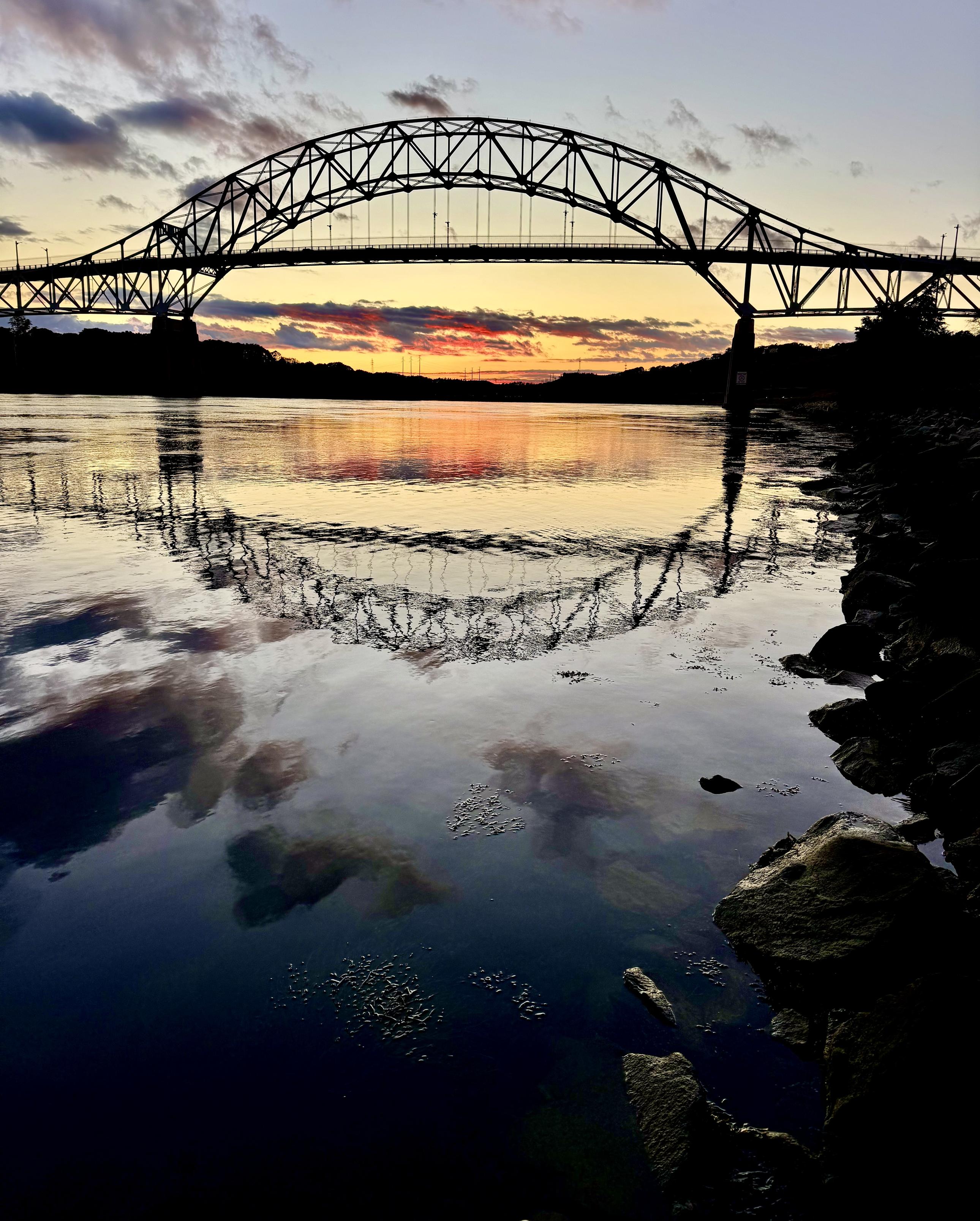 Photo of bridge over calm reflective water. Sunset in background. Dark clouds overhead and in reflections.
Sagamore Bridge, Massachusetts.
