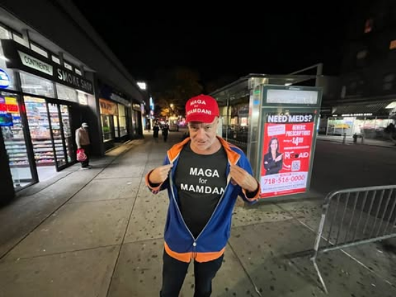 55 year old white man in NYC standing on a sidewalk wearing a red hat that says "MAGA for MAMDANI" and a black T shirt that says the same.