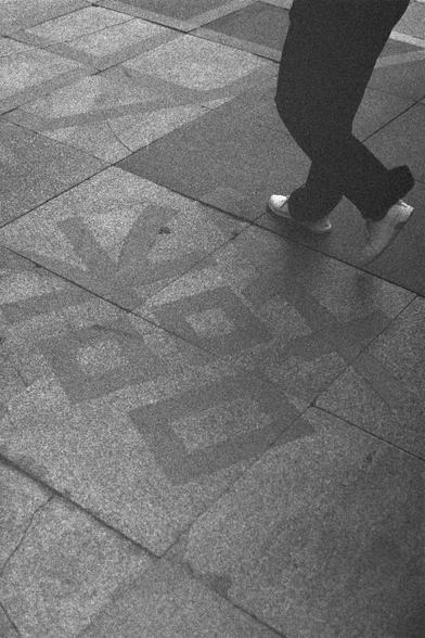 Ilford HP5 Plus 400 (FF)

English Alt Text:
A black-and-white image showing a person walking across a tiled pavement. Only the lower legs and feet are visible; the person wears dark pants and white shoes. The pavement features geometric patterns, including arrows and diamond shapes, embedded into the tiles. The image has a grainy texture, giving it an artistic, vintage appearance. The scene captures motion and design in an urban setting.

中文替代文本：
一张黑白照片，展示一个人走在铺有几何图案的地砖上。画面中只看到其腿部和脚部，穿着深色裤子和白色鞋…