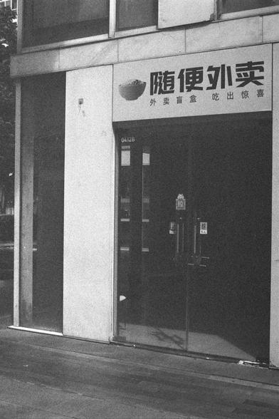 Ilford HP5 Plus 400 (FF)

English Alt Text:
A storefront entrance with glass doors and signage above. The sign includes Chinese text and an image of a bowl, suggesting a food establishment. The text reads “随便外卖 外卖盲盒 吃出惊喜,” promoting takeout meals with a surprise element. A unit number “0412b” is visible above the door. The setting is urban, with reflections of buildings in the glass. The image is in black and white, giving it a stylized, vintage feel.

中文替代文本：
一家餐饮店的入口，设有玻璃门和上方招牌。招牌上有中文文字和一个碗的图…