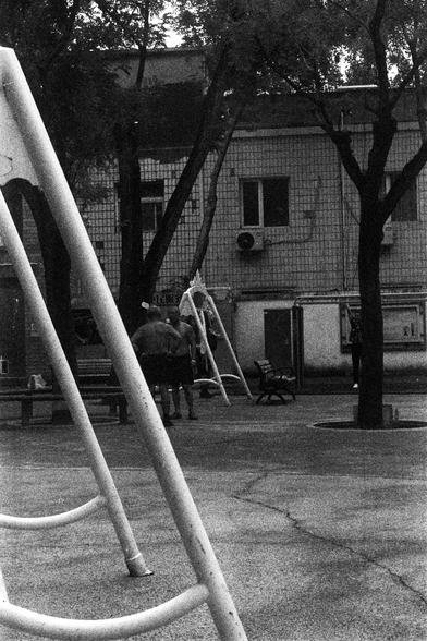 ORWO P400 (FF)

English Alt Text: A black-and-white photo of a playground in an urban setting. In the foreground, a large metal climbing frame with curved bars dominates the scene. In the background, people gather near another play structure, possibly a slide. The ground is paved, and trees offer shade. A building with tiled walls, windows, and air conditioning units stands behind the playground. The image has a grainy texture, evoking a vintage or film-like quality.

中文替代文字：
这是一张黑白照片，拍摄于城市中的一个…