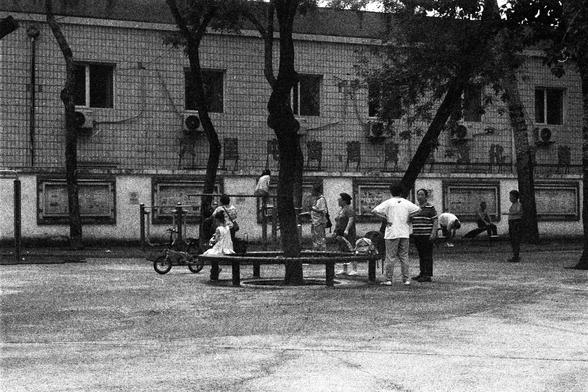 ORWO P400 (FF)

English Alt Text: A grayscale photo of a public courtyard or park. A circular bench surrounds a tree in the center, with people standing, sitting, stretching, and chatting nearby. The setting is lively and communal. A scooter is parked on the left. In the background, a multi-story building with tiled walls, windows, and air conditioning units frames the scene. Trees are scattered throughout, adding shade and texture to the space.

中文替代文字：
一张灰度照片，展示一个公共庭院或公园。画面中央是一棵树，周围环绕着圆形长椅。人们…
